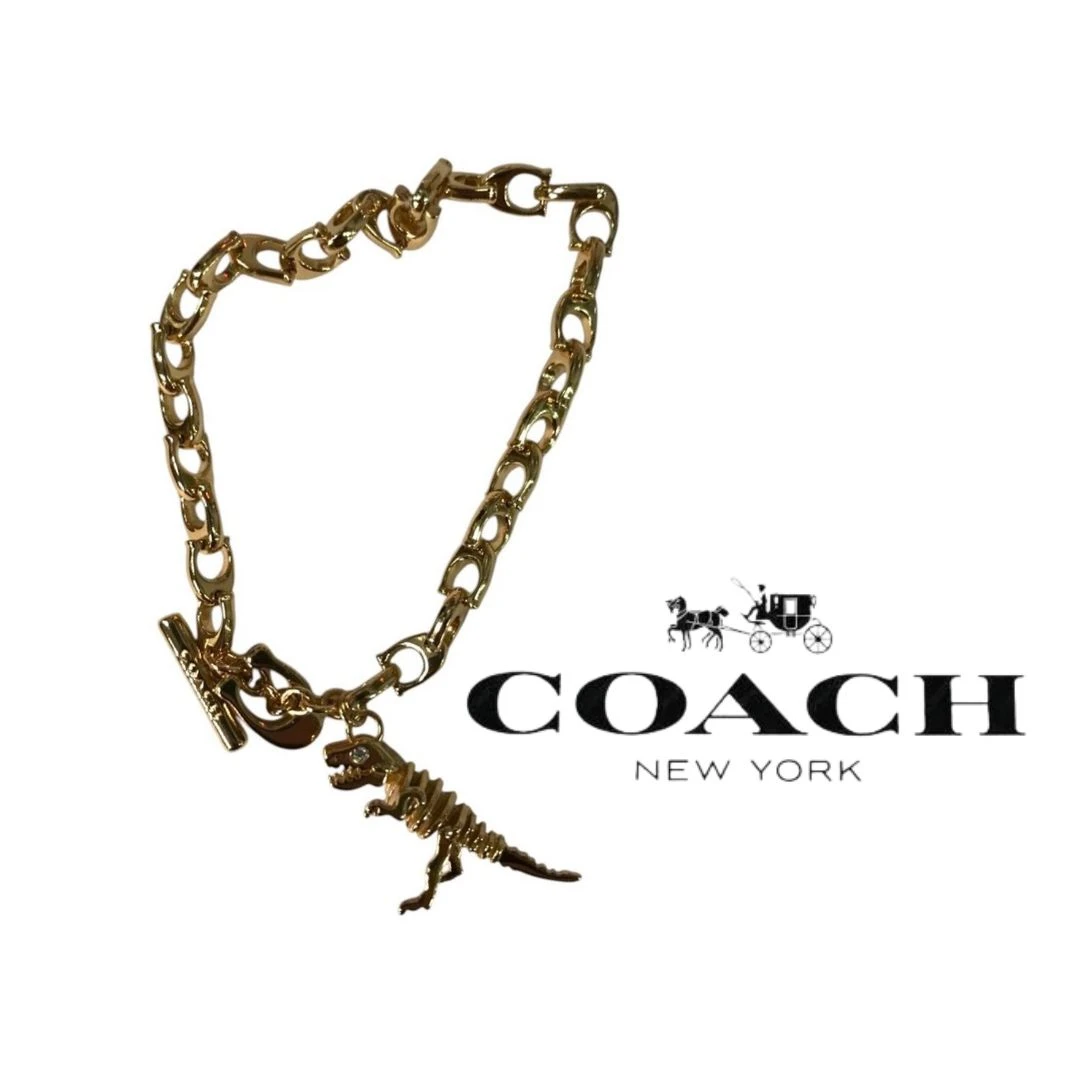 PULSERA COACH SKELETAL REXY CON DIJE DE DINOSAURIO – TONO DORADO NUEVO.